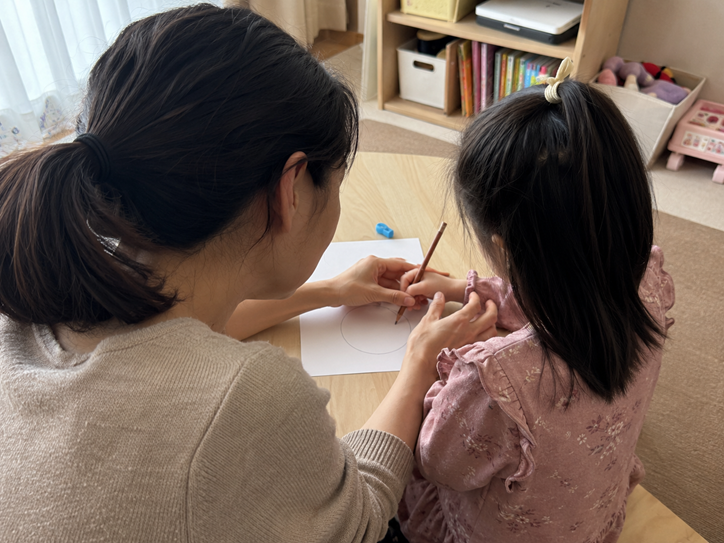 A mother gently guides her young daughter’s hand and pencil grip while she practices drawing a simple shape.
