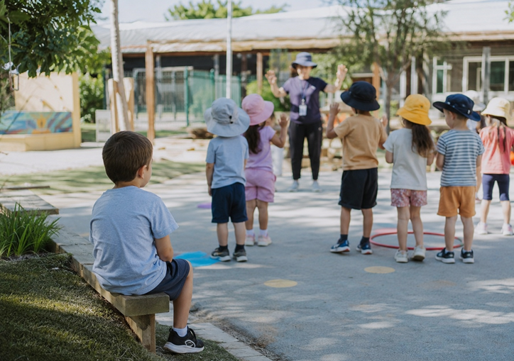 Child with ASD level 1 diagnosis sits slightly apart while other children take part in structured playground play, reflecting subtle social challenges associated with ASD Level 1.